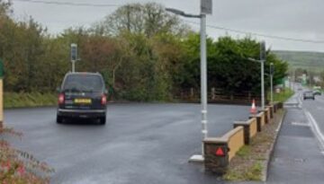 DBS805 lighting car park, A dark van is parked on a wet asphalt surface near a row of solar-powered streetlights and a low stone wall bordering a road.