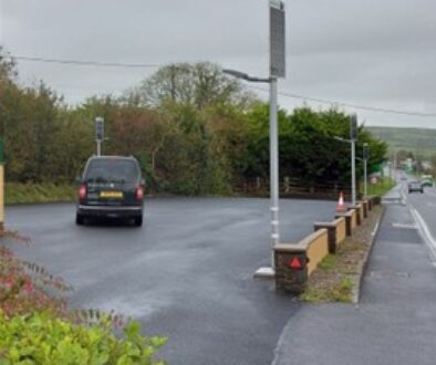 DBS805 lighting car park, A dark van is parked on a wet asphalt surface near a row of solar-powered streetlights and a low stone wall bordering a road.
