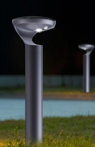 Solar bollard ground mounted lighting system. A modern, dark gray bollard light with a solar panel and upward-facing LED light illuminates a grassy area next to a body of water at night, with a second, bollard light visible in the background.
